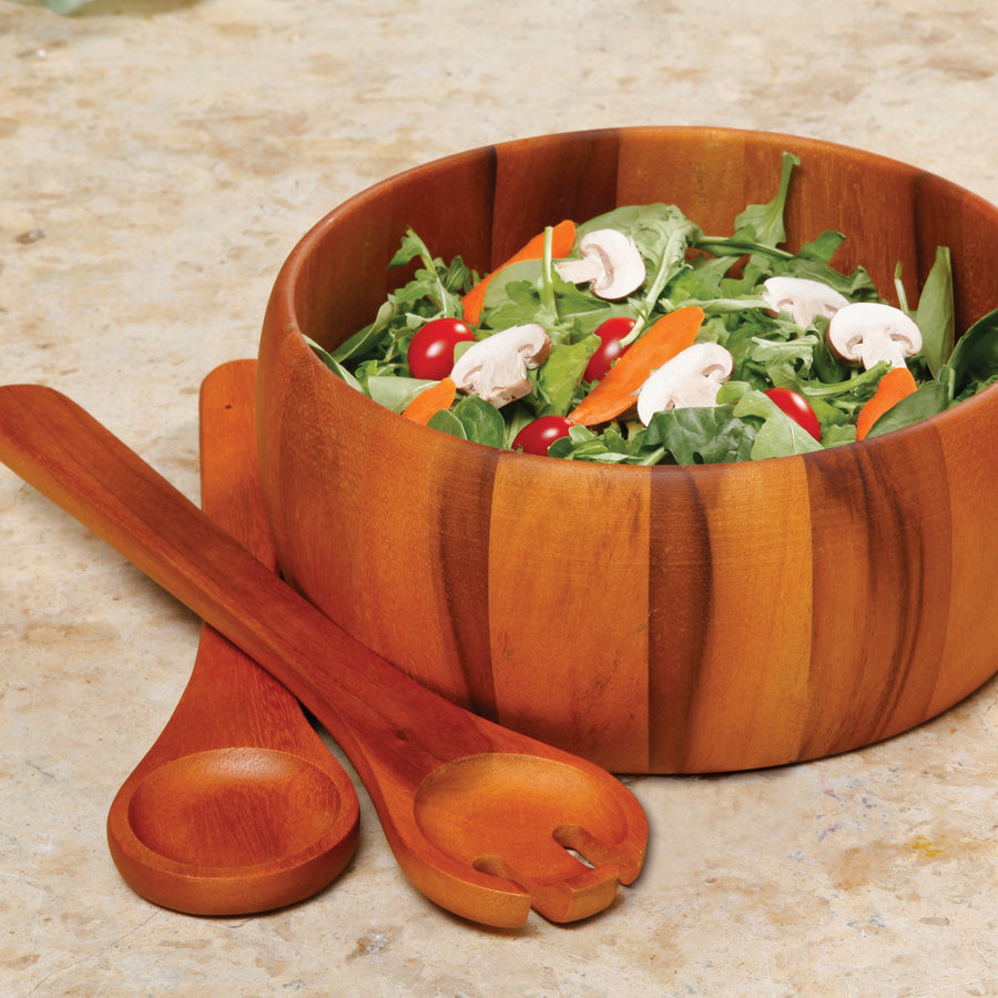 Laroda acacia wood salad bowl with mixed greens, mushrooms and tomatoes, serving spoon and fork resting beside it on stone