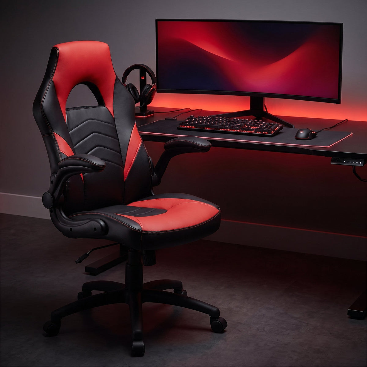 Black and red gaming chair with flip-up arms at a battle station desk with a red-lit keyboard, monitor and headset at night