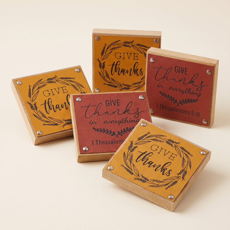 Six small square harvest signs in yellow, red and brown reading give thanks and be thankful, raised studio angle on a plain surface