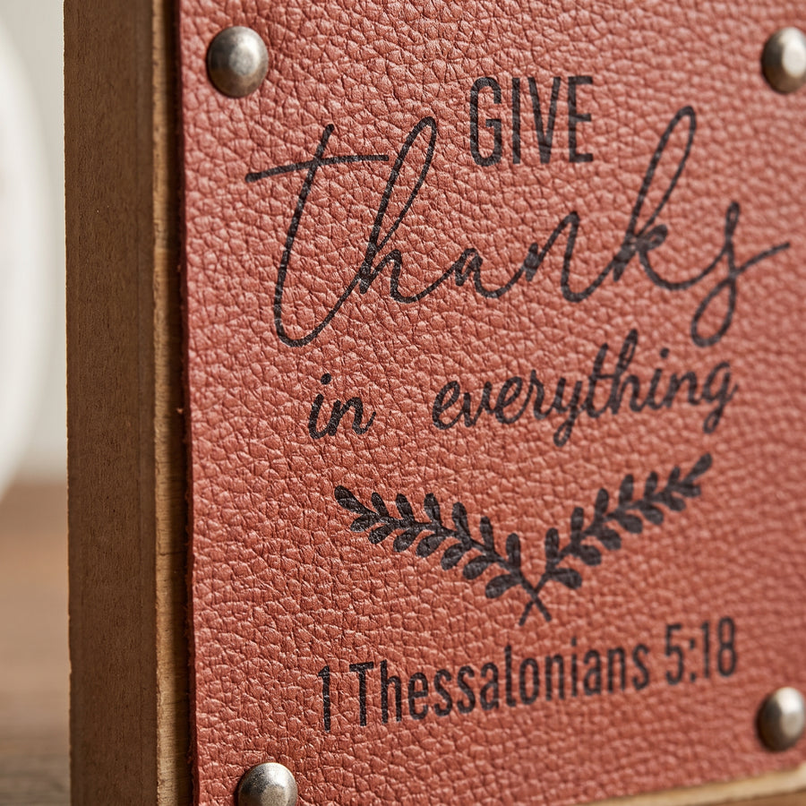 Macro of a small harvest sign showing wood face, faux leather accent and printed give thanks wording