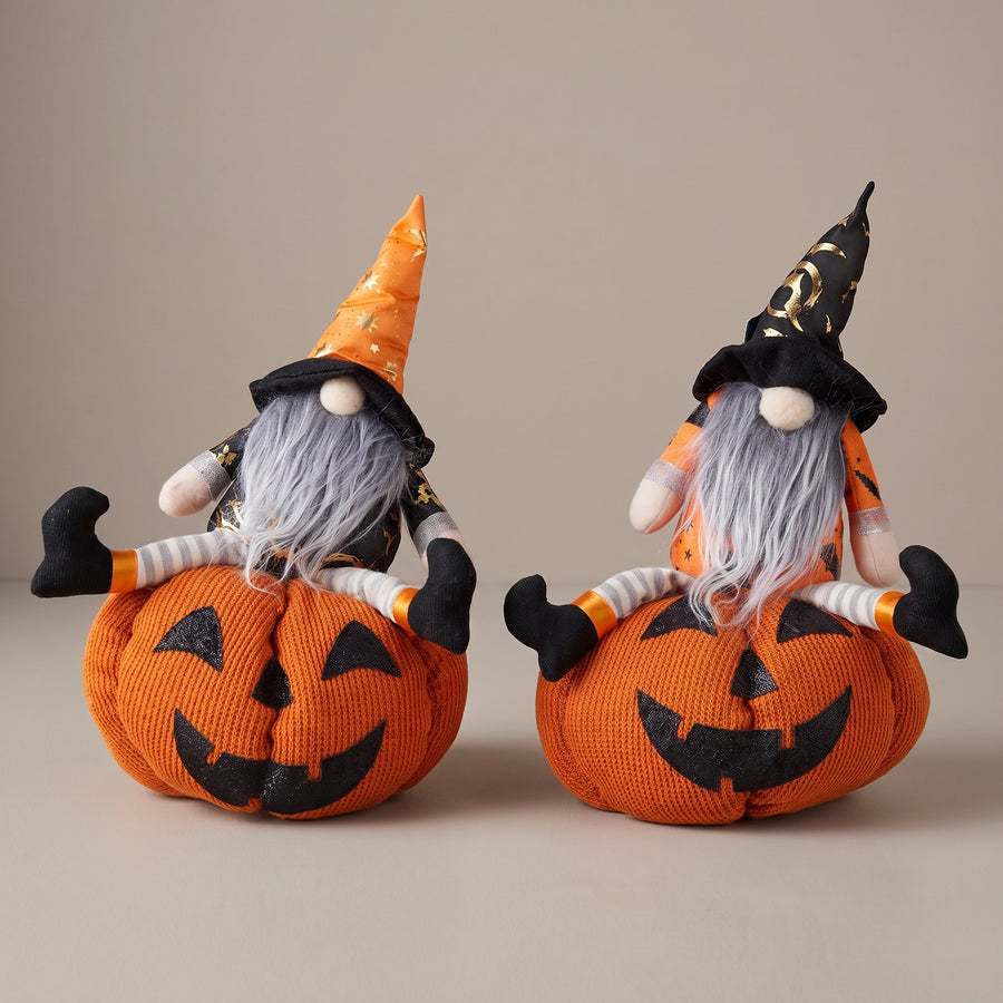 Two plush gnomes seated on bright orange jack-o-lantern pumpkins, about fourteen inches, raised studio angle on a plain surface
