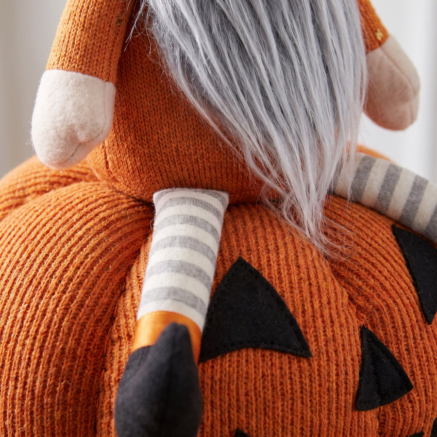Macro of a plush gnome perched on an orange plush pumpkin showing fabric nap and stitched ribs