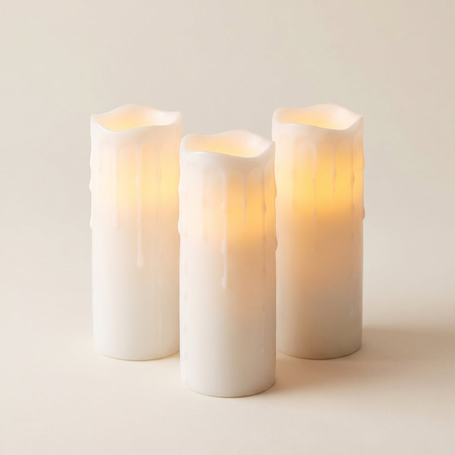 Three tall white dripping-wax LED pillar candles about three by eight inches, matched in height and glowing warmly from inside, raised studio angle on a plain surface