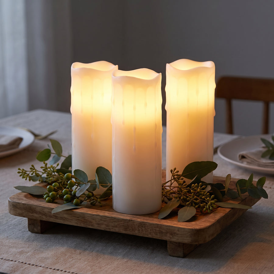 Three tall glowing white pillar candles clustered as a centerpiece on a dining table at dusk
