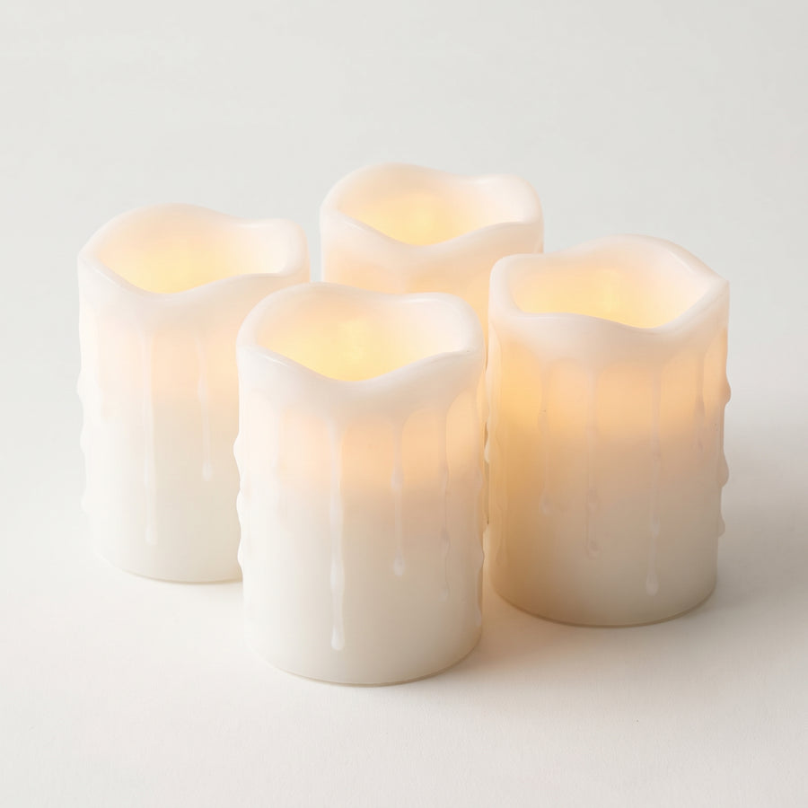 Four white dripping-wax LED pillar candles about three by four inches, matched in height and glowing warmly from inside, raised studio angle on a plain surface