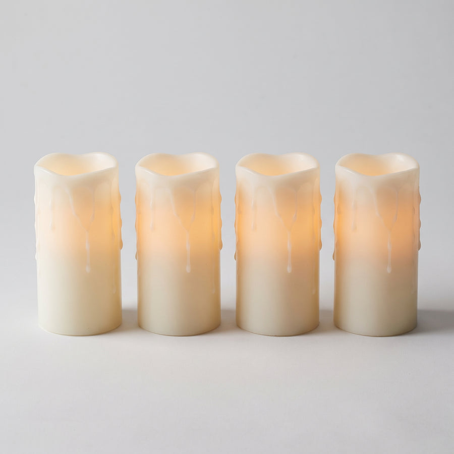 Four tall white dripping-wax LED pillar candles about three by six inches, matched in height and glowing warmly from inside, raised studio angle on a plain surface