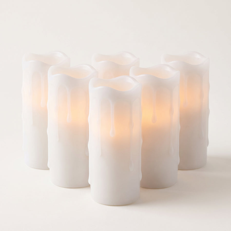 Six slim white dripping-wax LED pillar candles about one and three quarter by four inches, matched in height and glowing warmly from inside, raised studio angle on a plain surface