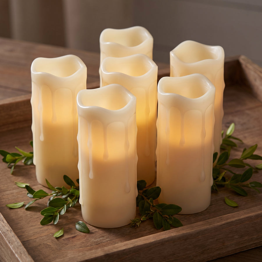 Six slim glowing white pillar candles clustered on a mirrored tray at dusk with greenery