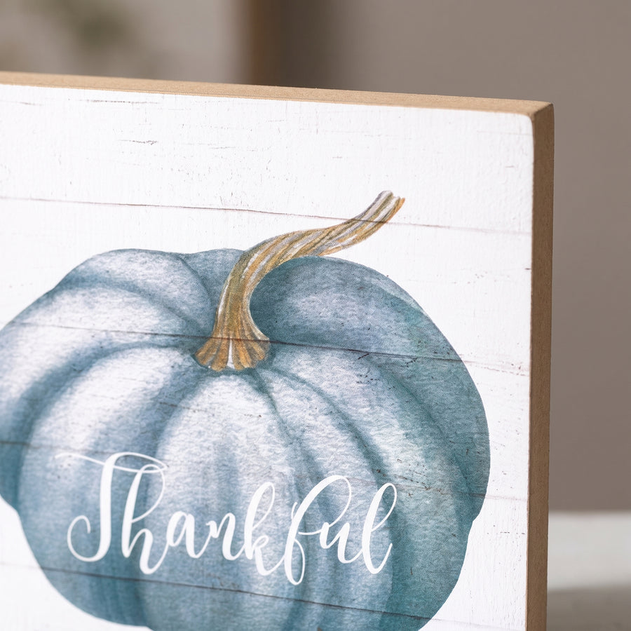 Macro of a square white shiplap sign showing a printed blue watercolour pumpkin and the word thankful in white script