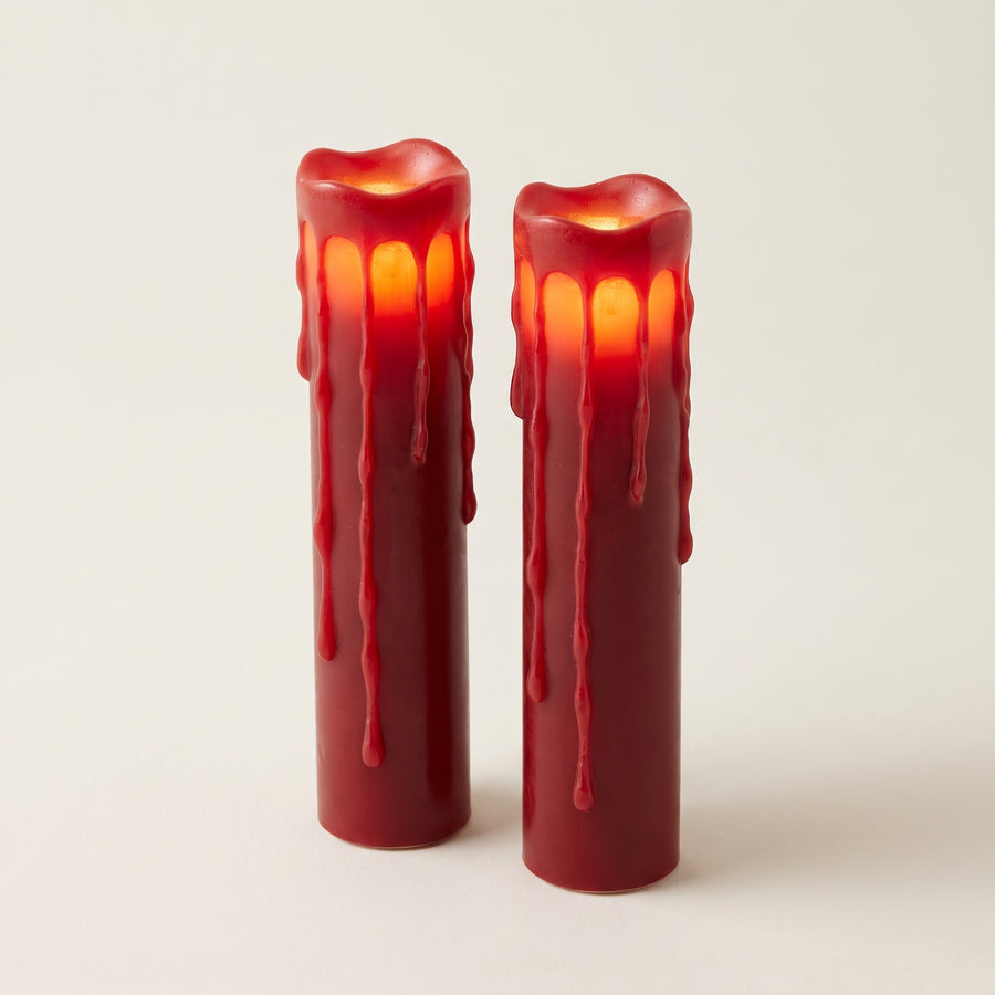 Two tall red dripping-wax LED pillar candles about two by eight inches, matched in height and glowing warmly from inside, raised studio angle on a plain surface
