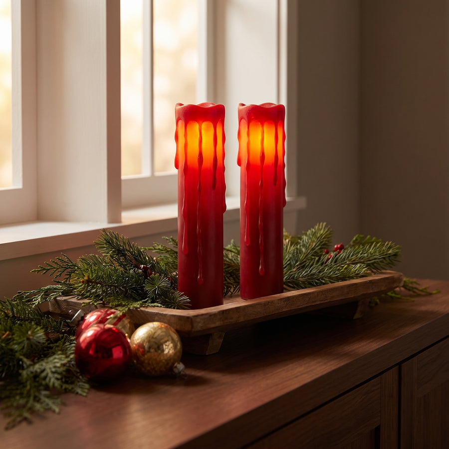 Two tall glowing red pillar candles on a holiday sideboard at dusk with evergreen