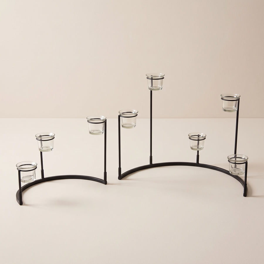 Two black metal crescent votive stands, one holding three glass cups and one holding four, raised studio angle on a plain surface