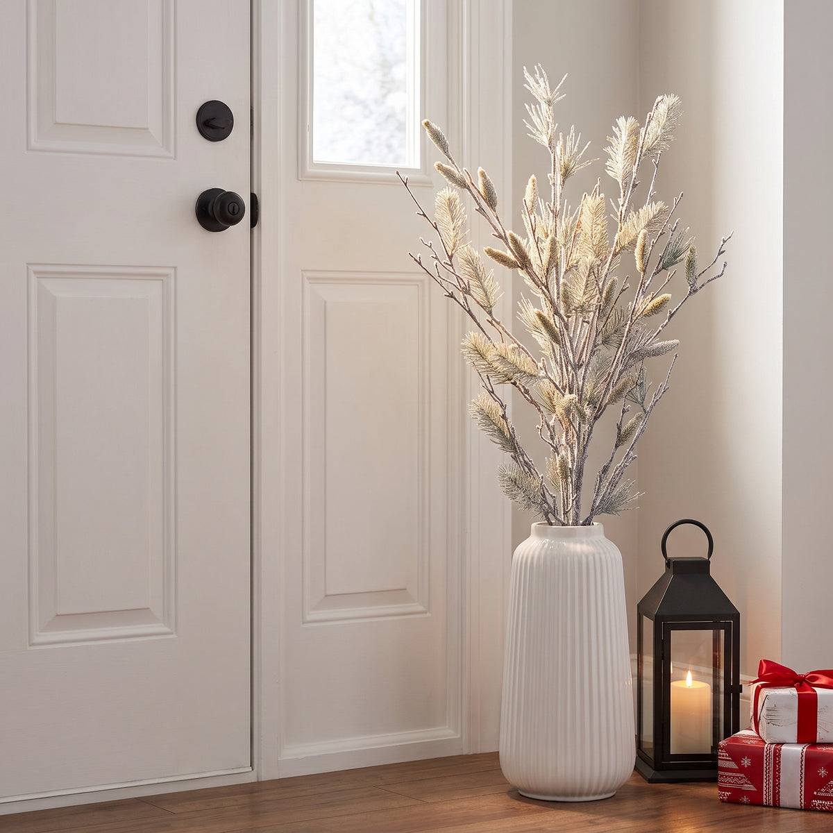 Oversized snowy pine bundle in a tall white floor vase against an entryway wall beside a lantern