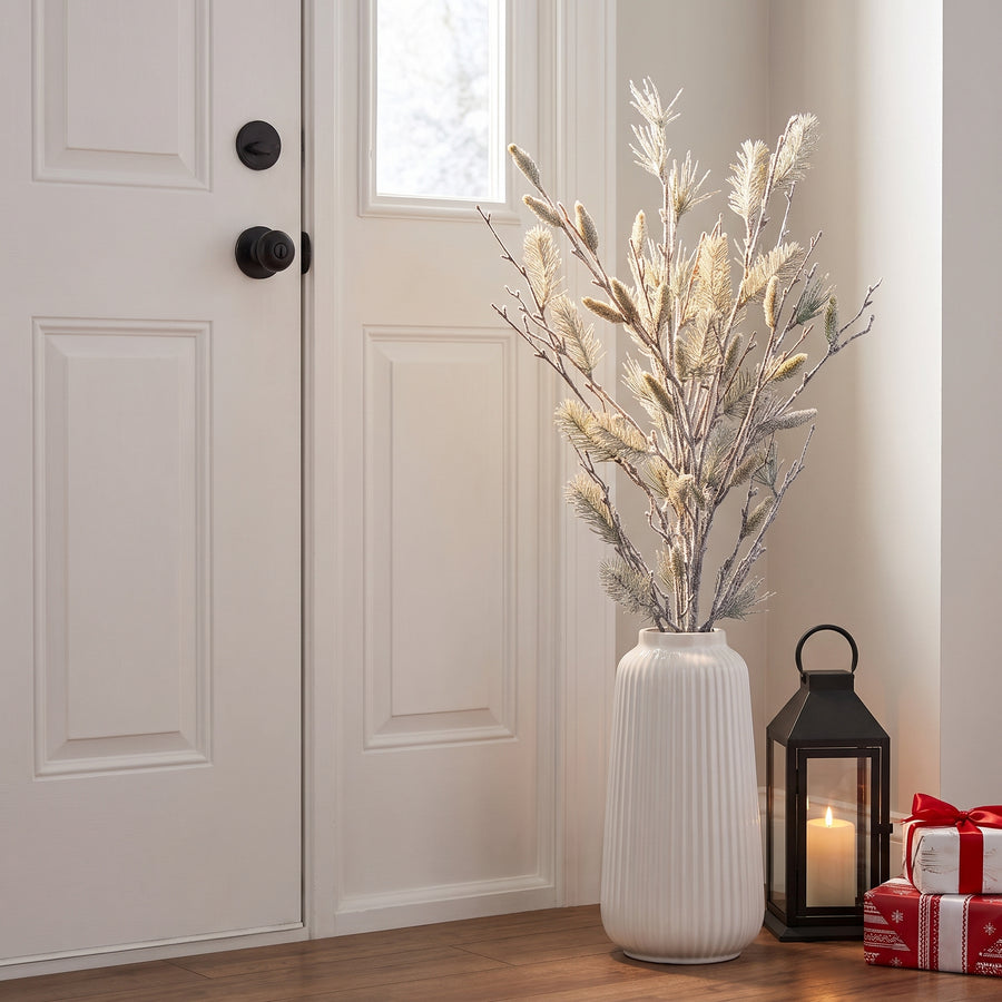 Oversized snowy pine bundle in a tall white floor vase against an entryway wall beside a lantern