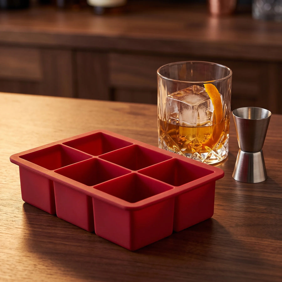 Red silicone large cube ice tray beside a whiskey glass with one big square ice cube on a walnut bar top, Houdini
