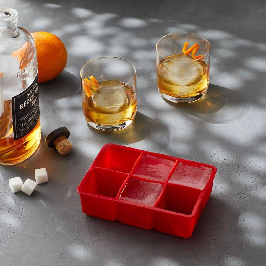 Red silicone ice cube tray next to whiskey glasses with large clear ice cubes, an orange peel and sugar cubes